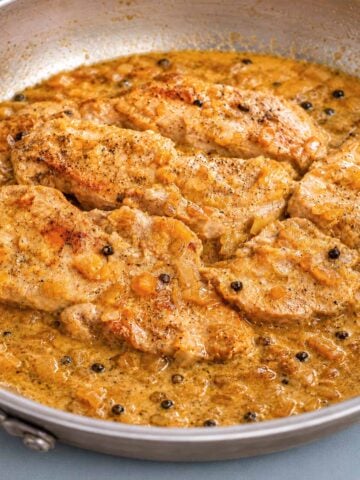 Pork chops in peppercorn sauce cooking in a skillet with onions and whole black peppercorns