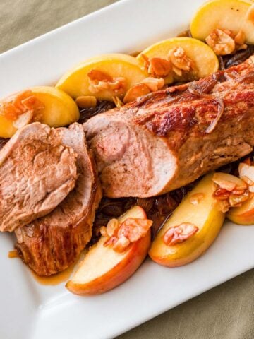 Pork tenderloin sliced and served on a white platter with caramelized apple wedges and pan sauce