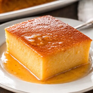 Pumpkin flan made with condensed milk, served as a thick slice with a glossy caramel top and caramel sauce pooling on a white plate, with the remaining flan in the background.