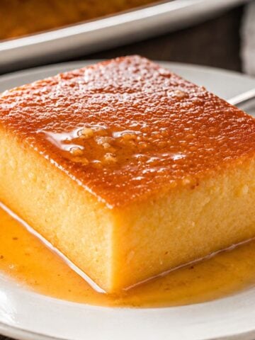Pumpkin flan made with condensed milk, served as a thick slice with a glossy caramel top and caramel sauce pooling on a white plate, with the remaining flan in the background.