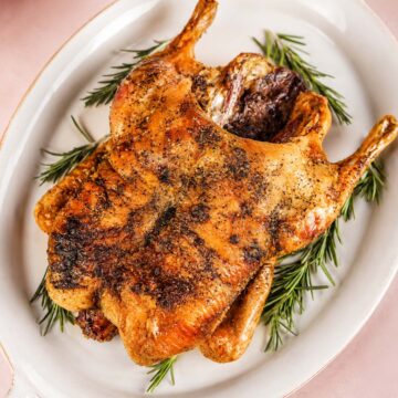 Whole roasted duck with crisp, golden-brown skin and black pepper seasoning, served on a white platter with fresh rosemary sprigs.