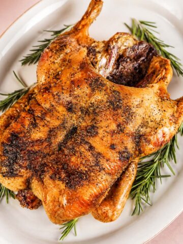 Whole roasted duck with crisp, golden-brown skin and black pepper seasoning, served on a white platter with fresh rosemary sprigs.
