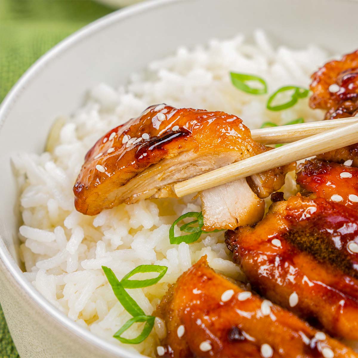 Chopsticks holding a piece of teriyaki chicken over white rice, garnished with sesame seeds and sliced green onions.