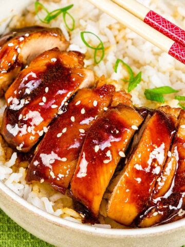 Teriyaki chicken sliced over white rice in a bowl, garnished with sesame seeds and green onions, served with chopsticks.
