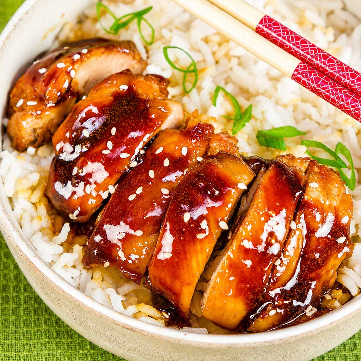 Teriyaki chicken sliced over white rice in a bowl, garnished with sesame seeds and green onions, served with chopsticks.