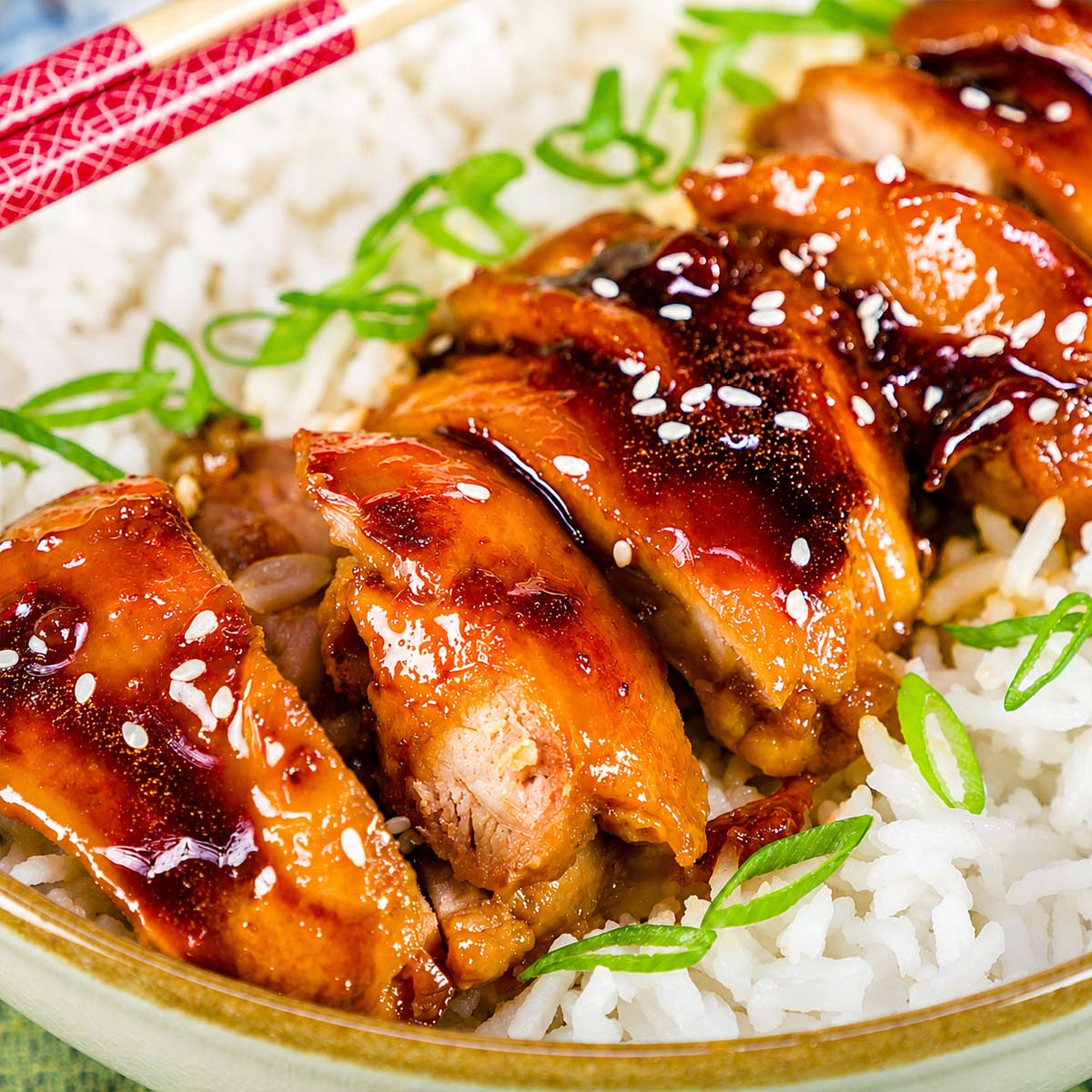 Teriyaki chicken sliced over white rice in a bowl, garnished with sesame seeds and green onions, served with chopsticks.
