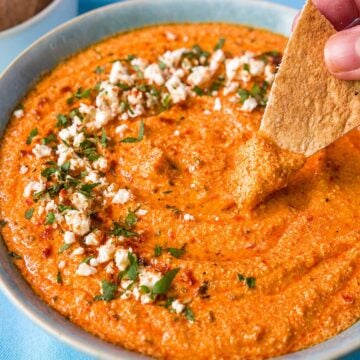 Pita chip dipping into creamy tirokafteri Greek spicy feta dip garnished with crumbled feta, parsley, and olive oil.