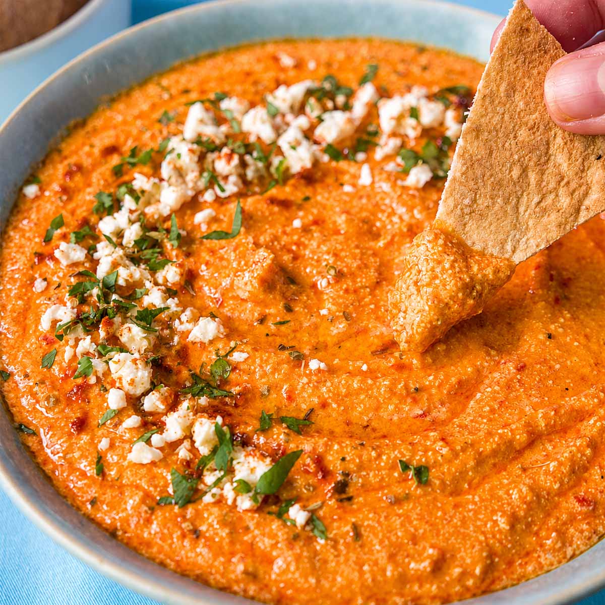 Pita chip dipping into creamy tirokafteri Greek spicy feta dip garnished with crumbled feta, parsley, and olive oil.