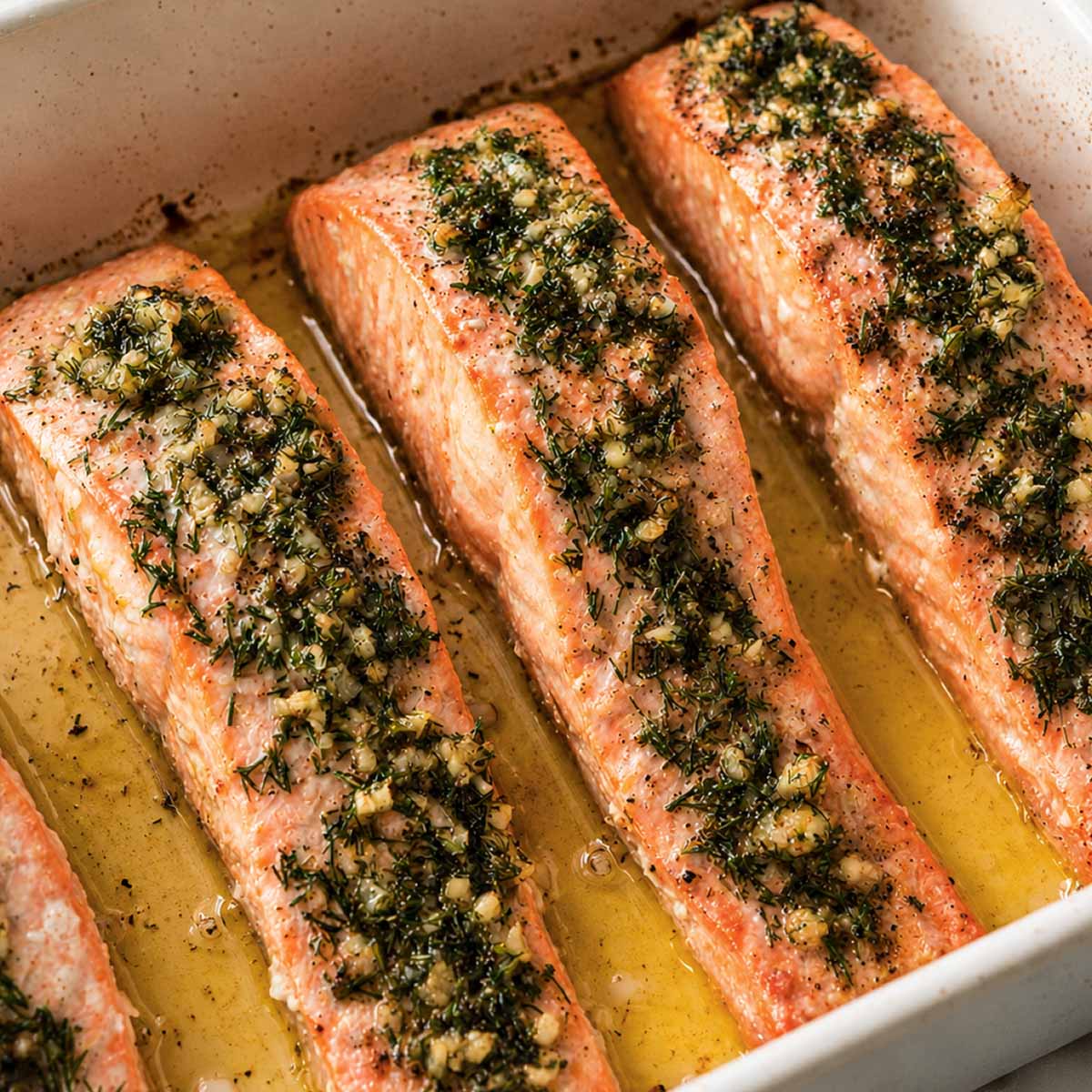 close up baked lemon dill salmon fillets in a white baking dish with garlic, fresh dill, and olive oil juices