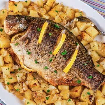 Baked whole tilapia with lemon slices on top, served on a white platter over roasted potatoes and onions
