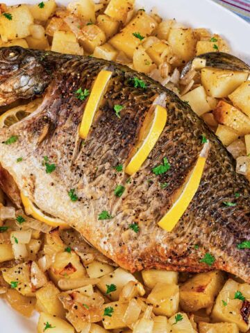 Baked whole tilapia with lemon slices on top, served on a white platter over roasted potatoes and onions