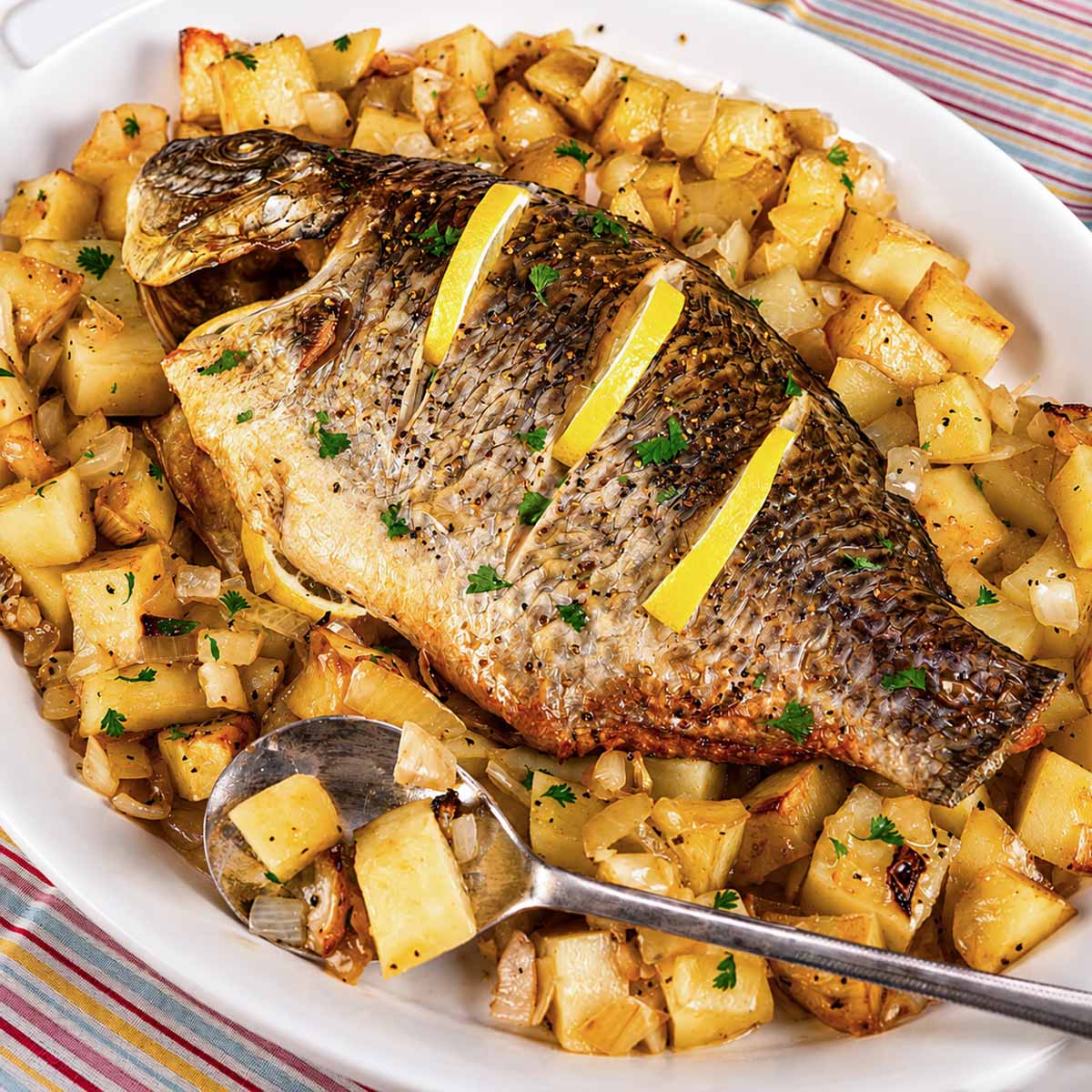 Baked whole tilapia with lemon slices on top, served on a white platter over roasted potatoes and onions.