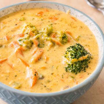 Close-up of creamy broccoli cheddar soup with melted cheddar, broccoli florets, and grated carrot in a patterned bowl.