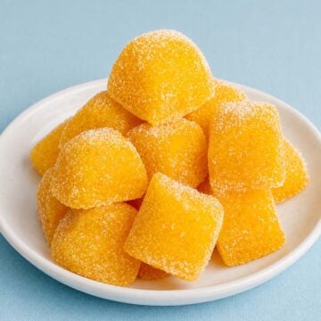 Homemade fruit snacks without gelatin, sugar-coated orange gummy squares stacked on a white plate with a hand picking one up.