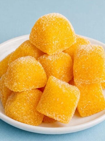 Homemade fruit snacks without gelatin, sugar-coated orange gummy squares stacked on a white plate with a hand picking one up.