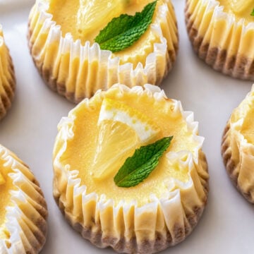 Mini lemon cheesecakes in paper liners topped with lemon slices and mint leaves on a tray.