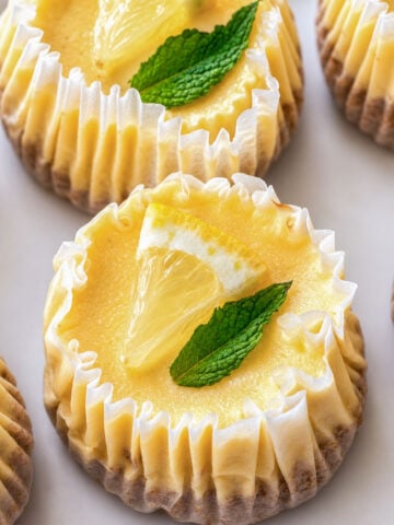 Mini lemon cheesecakes in paper liners topped with lemon slices and mint leaves on a tray.