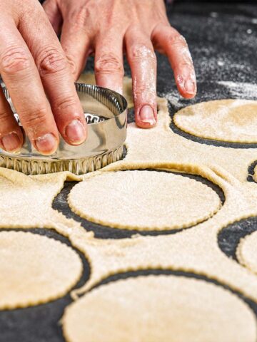 Hands using a round cutter to cut pierogi dough circles from rolled dough on a floured surface.