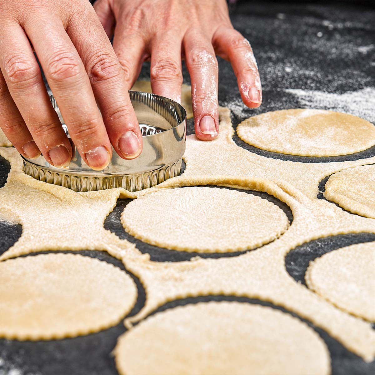 Hands using a round cutter to cut pierogi dough circles from rolled dough on a floured surface.
