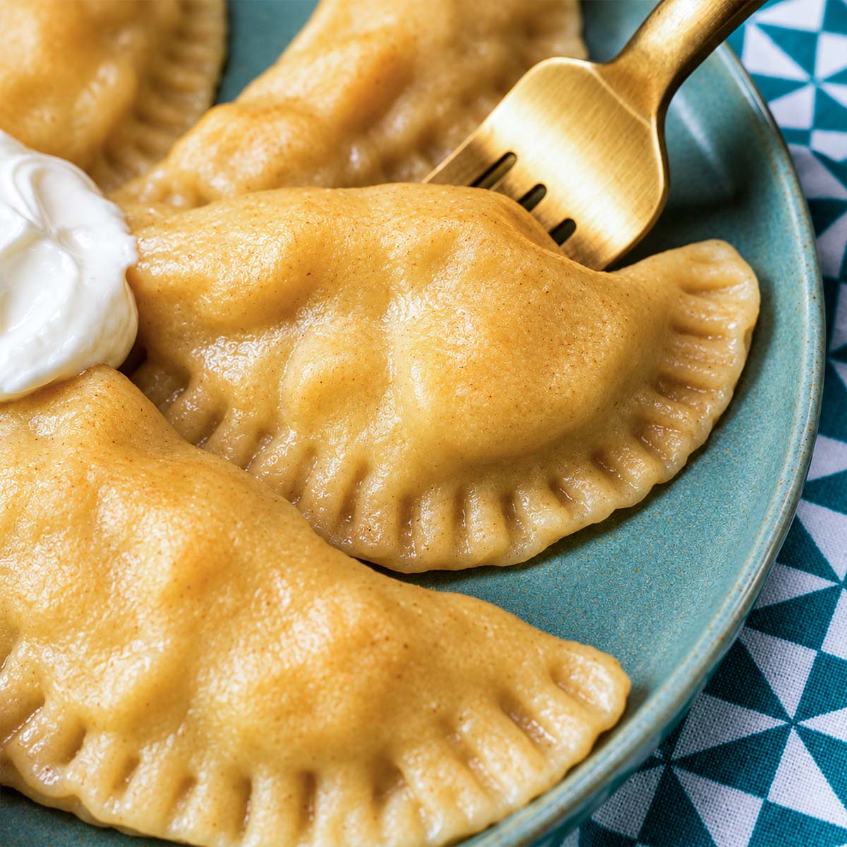 Cooked pierogi on a plate with crimped edges, served with a dollop of sour cream and a fork.