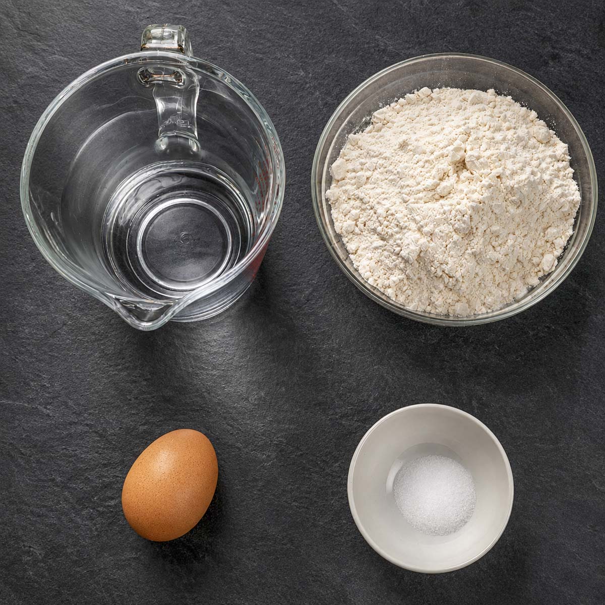Pierogi dough ingredients on a dark surface: flour in a bowl, water in a measuring jug, a whole egg, and salt in a small dish.