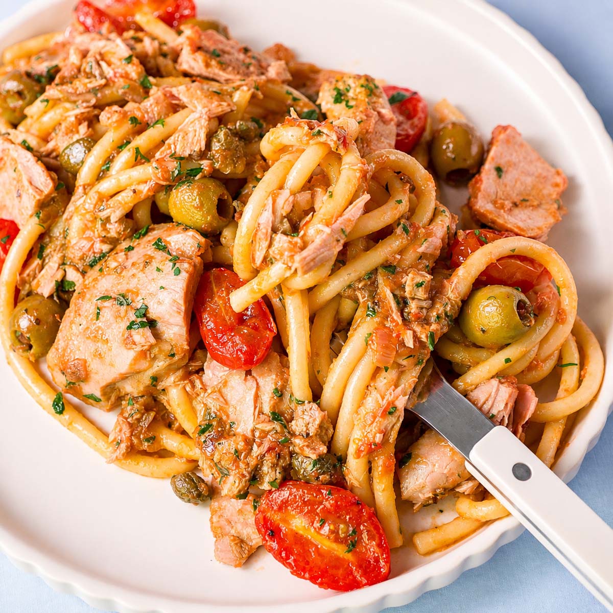 Fork lifting tuna pasta with tomatoes, green olives, capers, and parsley