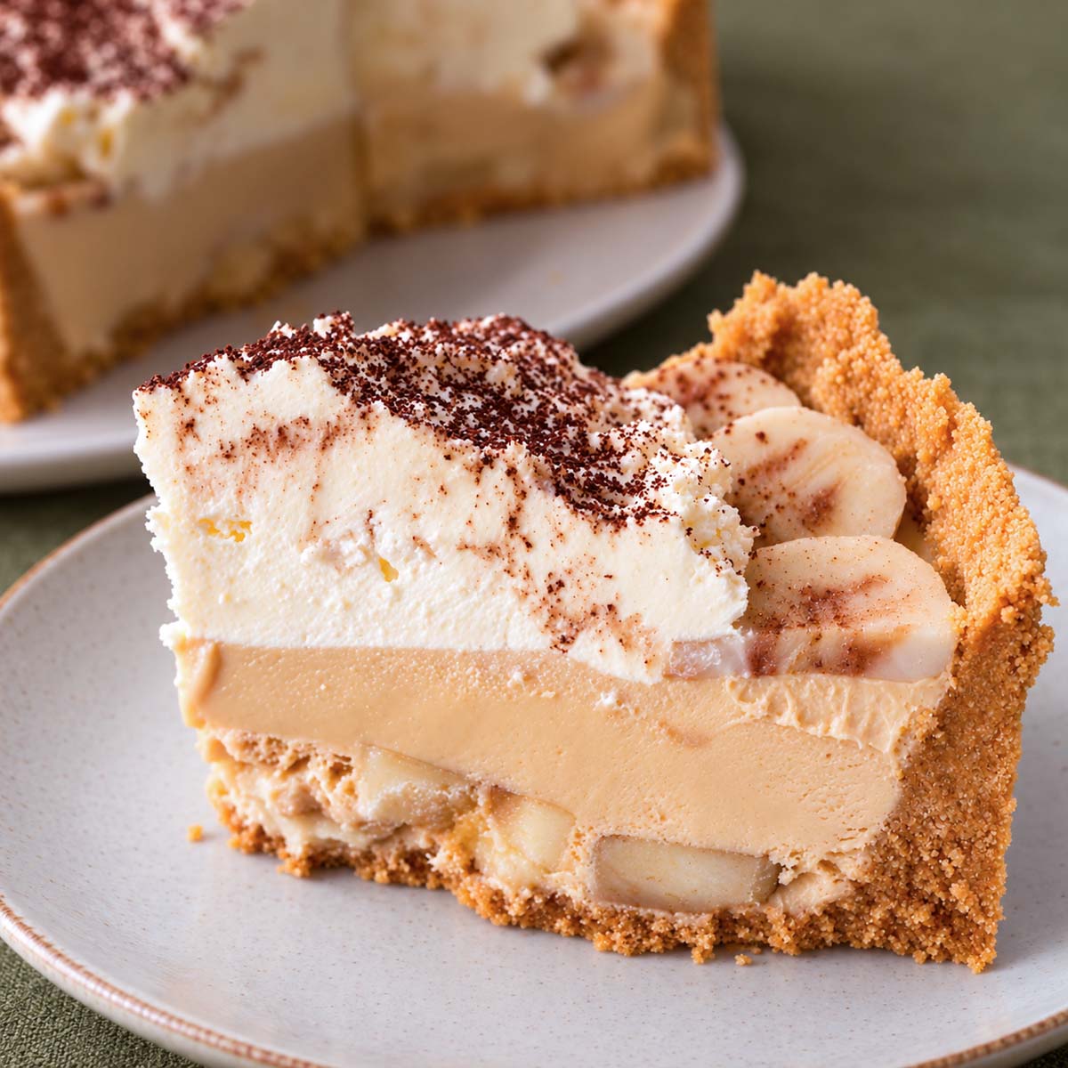 Slice of banoffee pie showing biscuit crust, banana, creamy caramel filling and whipped cream and chocolate dusting