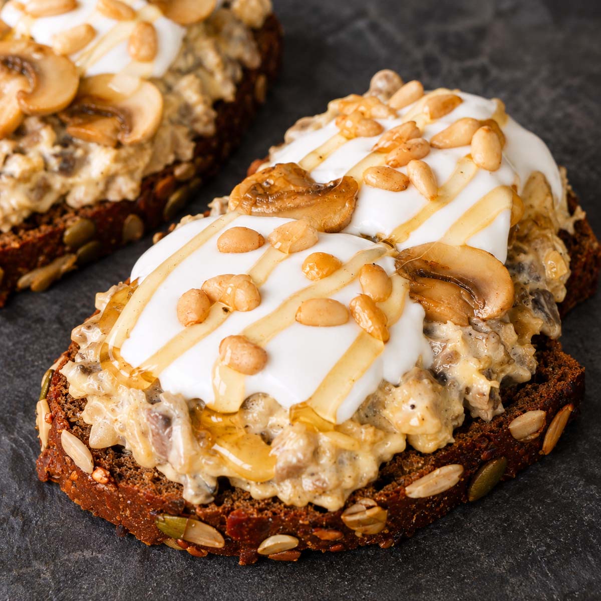 Creamy mushroom toast topped with garlic aioli, sautéed mushrooms, pine nuts, and a drizzle of honey on a dark board.