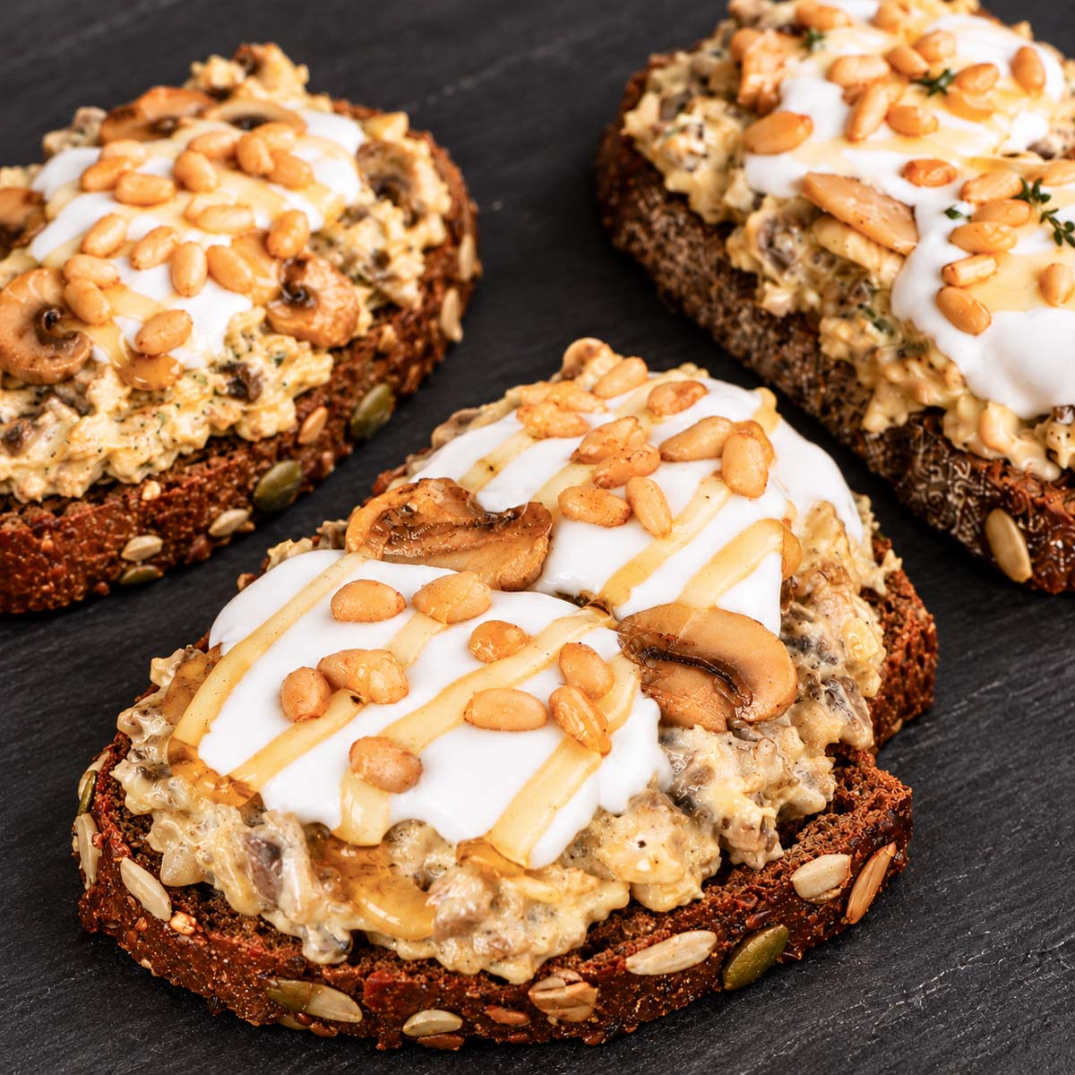 Three creamy mushroom toasts topped with garlic aioli, pine nuts, sautéed mushrooms, and honey on dark slate.