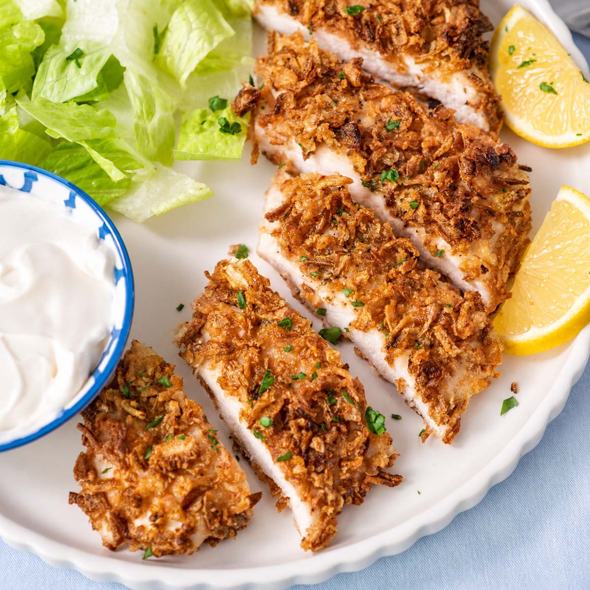 Sliced crispy French onion chicken showing the white meat inside, served with lettuce, lemon wedges, and creamy dipping sauce.