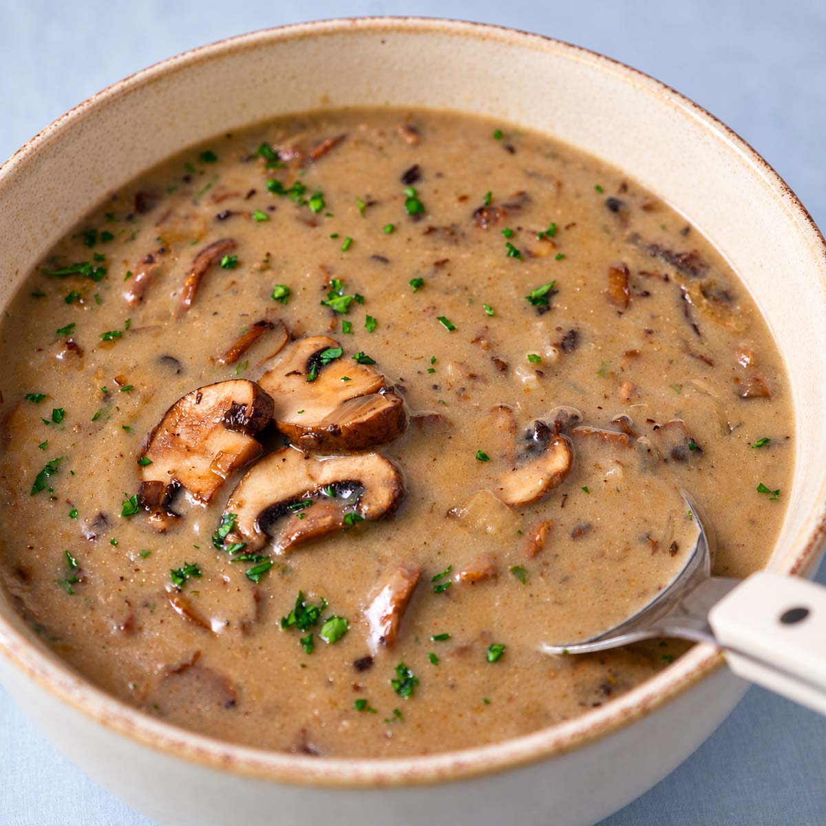Bowl of mushroom soup without cream with sliced mushrooms, herbs, and a spoon.