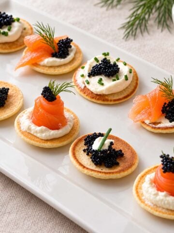 Smoked salmon blinis topped with crème fraîche, black fish roe, dill, and chives on a white serving platter.