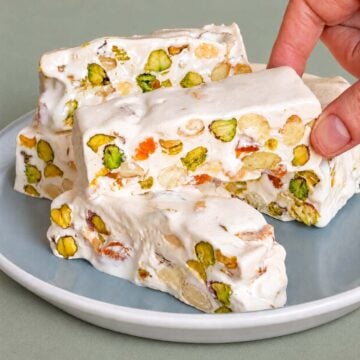 Hand holding a piece of pistachio nougat over a plate of nougat bars, showing creamy white nougat packed with pistachios and small bits of dried fruit.