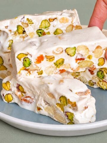 Hand holding a piece of pistachio nougat over a plate of nougat bars, showing creamy white nougat packed with pistachios and small bits of dried fruit.