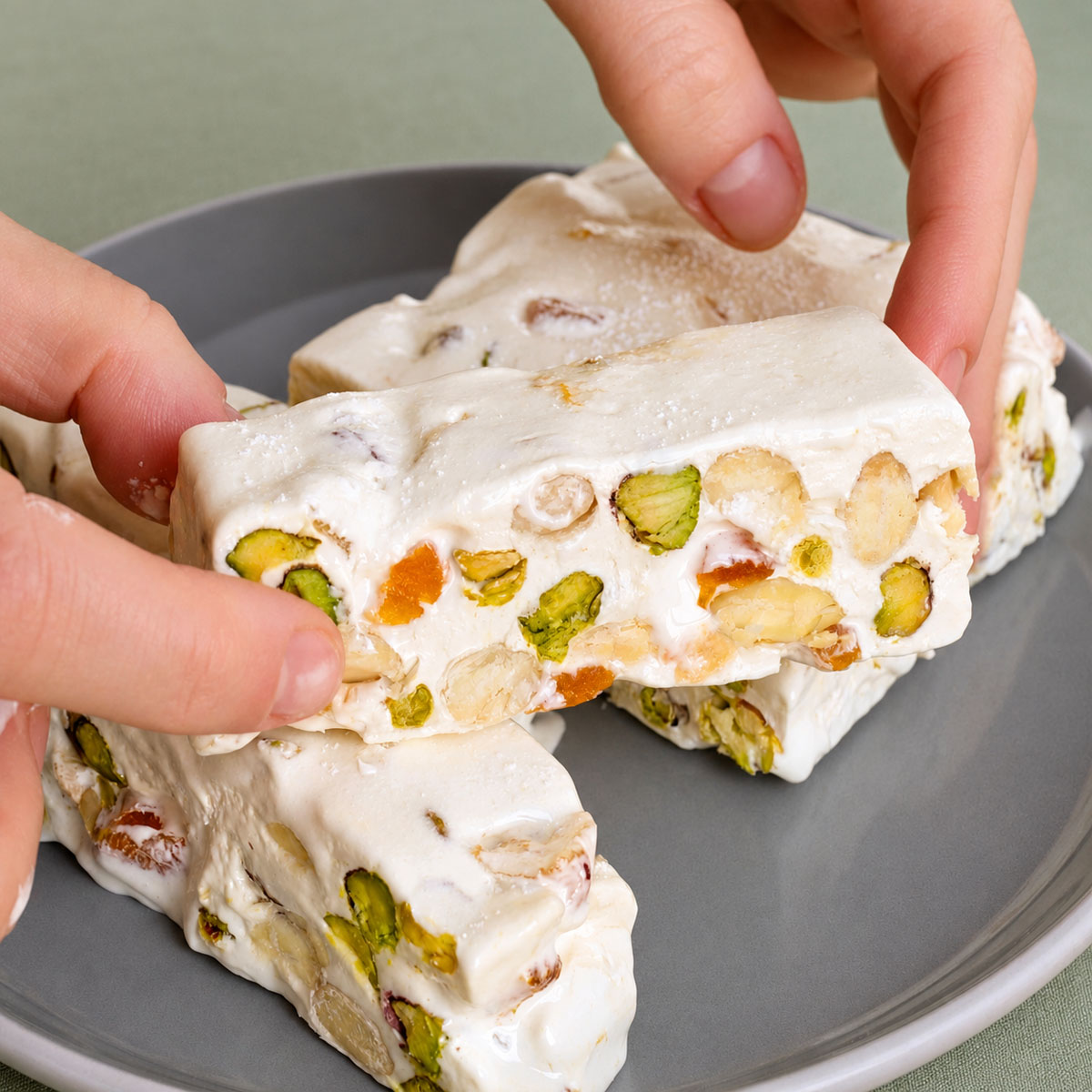 Hands arranging pistachio nougat bars on a plate, showing white nougat filled with pistachios, almonds, and bits of dried apricot. 