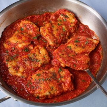 Boneless, skinless chicken thighs in roasted red pepper sauce, topped with chopped parsley in a stainless steel skillet.