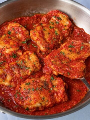 Boneless, skinless chicken thighs in roasted red pepper sauce, topped with chopped parsley in a stainless steel skillet.