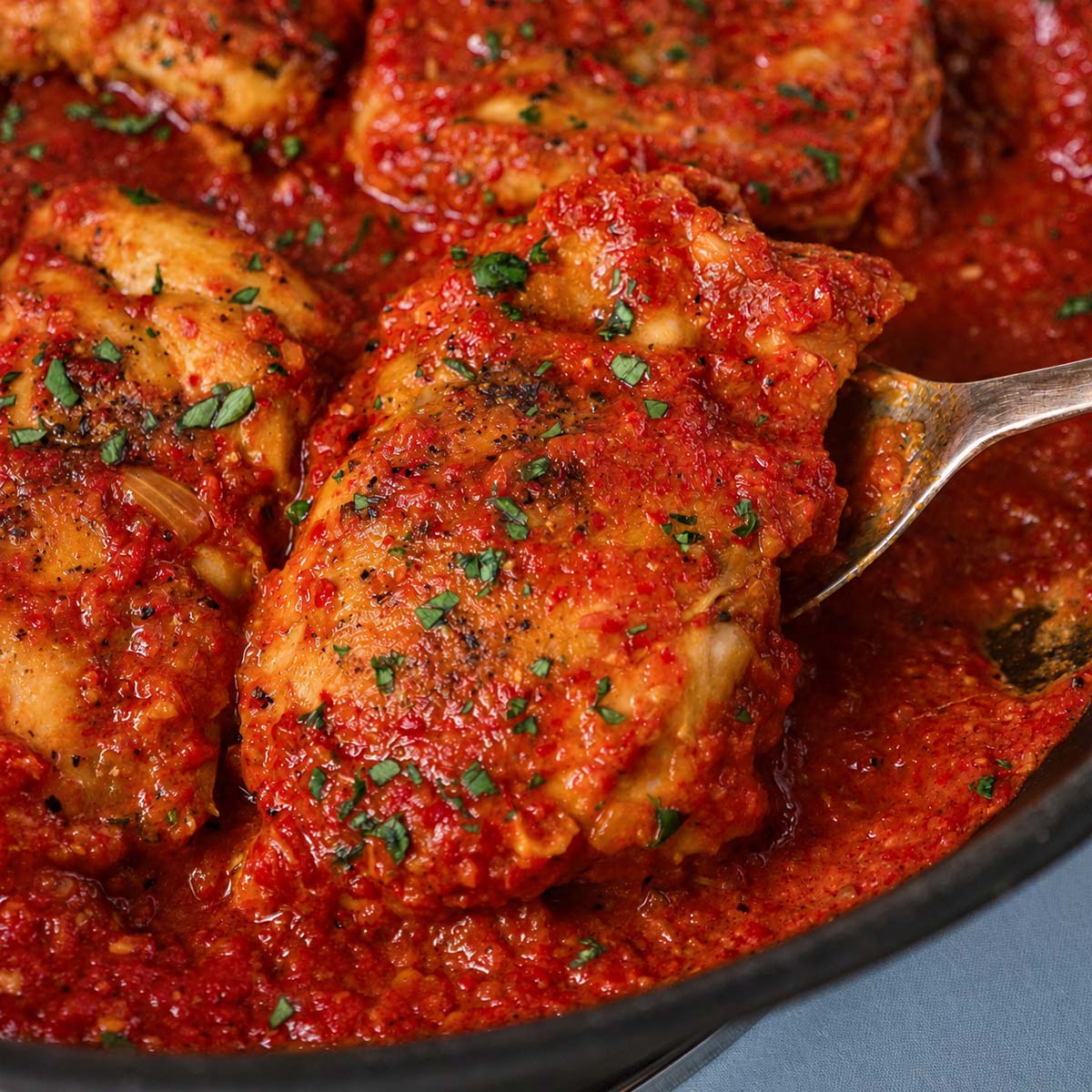 A chicken thigh lifted on a serving spoon from a stainless steel skillet of roasted red pepper sauce and parsley.