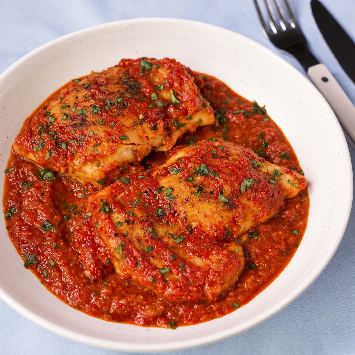 Two chicken thighs covered in roasted red pepper sauce and chopped parsley on a white ceramic plate.