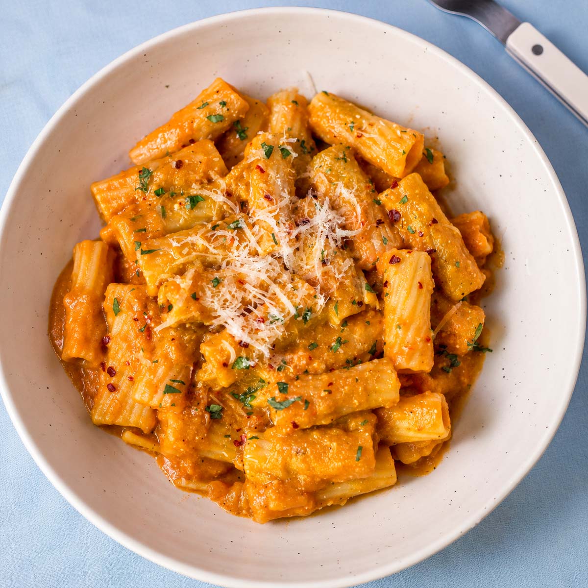 Creamy pumpkin rigatoni served in a white bowl with grated Parmesan, chopped parsley, and red pepper flakes.